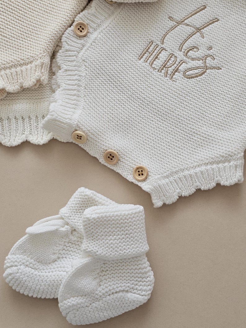 Puede incluir: Un conjunto de ropa de beb&eacute; de punto blanco, que incluye un pelele con botones de madera y el texto bordado "He's HERE", y patucos a juego. El pelele tiene un borde festoneado. El conjunto est&aacute; hecho de tejido de punto suave y texturizado.