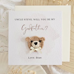 May include: A square greeting card with the text "UNCLE STEVE, WILL YOU BE MY Godfather?" and "Love, Rian." A crocheted teddy bear head with a white bow is centered on the card. The card is white with a textured surface.