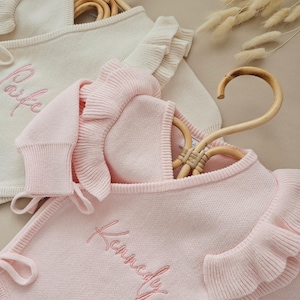 May include: Two baby outfits, one cream and one pink, displayed on wooden hangers. The cream outfit has the name "Paife" embroidered in pink. The pink outfit has the name "Kennedy" embroidered in pink. Both outfits have ruffled sleeves.