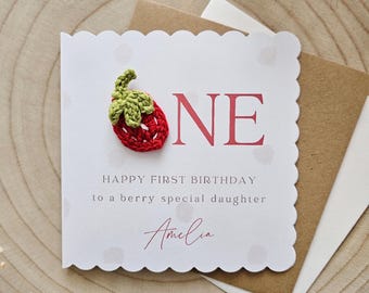 May include: A white card with a red crocheted strawberry and the word "ONE" in red letters. The card says "Happy First Birthday to a berry special daughter" and is signed "Amelia".