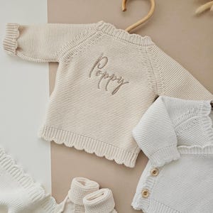 Puede incluir: Conjunto de ropa de beb&eacute; de punto color crema, que incluye un su&eacute;ter con el nombre "Poppy" bordado, una manta, patucos y una rebeca. El su&eacute;ter est&aacute; colgado en una percha de madera. Todos los art&iacute;culos son de color neutro.
