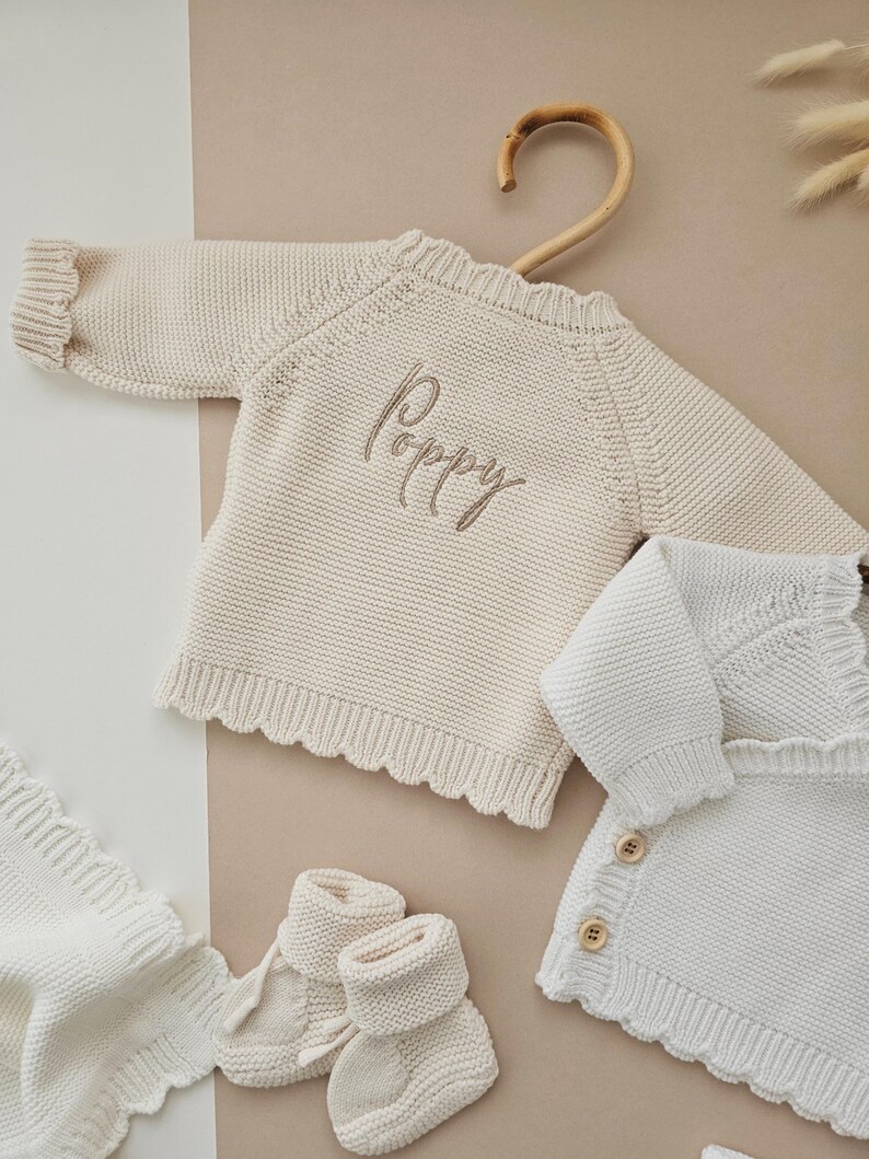 Puede incluir: Conjunto de ropa de beb&eacute; de punto color crema, que incluye un su&eacute;ter con el nombre "Poppy" bordado, una manta, patucos y una rebeca. El su&eacute;ter est&aacute; colgado en una percha de madera. Todos los art&iacute;culos son de color neutro.