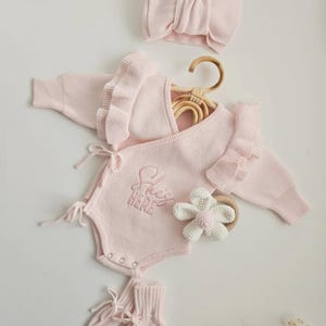 May include: A light pink baby clothing set, including a romper with the embroidered text "She's HERE", a headband, socks, and a flower-shaped teether. The romper has ruffle sleeves and tie closures. The set is displayed on a white surface.