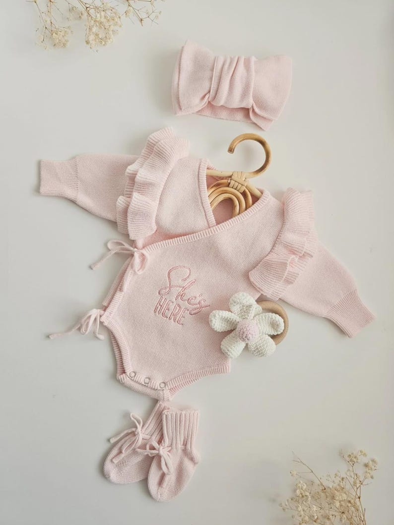 May include: A light pink baby clothing set, including a romper with the embroidered text "She's HERE", a headband, socks, and a flower-shaped teether. The romper has ruffle sleeves and tie closures. The set is displayed on a white surface.