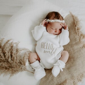 Puede incluir: Un beb&eacute; reci&eacute;n nacido con un body de punto blanco con las palabras "HELLO world" bordadas en la parte delantera. El beb&eacute; est&aacute; acostado sobre una almohada y una manta con flecos, con una diadema y calcetines a juego. Hierba de la pampa seca al lado.