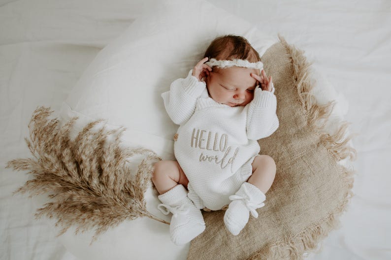 Puede incluir: Un beb&eacute; reci&eacute;n nacido con un body de punto blanco con las palabras "HELLO world" bordadas en la parte delantera. El beb&eacute; est&aacute; acostado sobre una almohada y una manta con flecos, con una diadema y calcetines a juego. Hierba de la pampa seca al lado.
