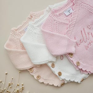 May include: Three knitted baby cardigans in light pink and white, featuring a scalloped edge, long sleeves, and wooden buttons along the side. The top pink cardigan displays the embroidered text "hello world" with a small heart icon.