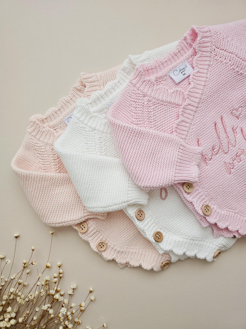 May include: Three knitted baby cardigans in light pink and white, featuring a scalloped edge, long sleeves, and wooden buttons along the side. The top pink cardigan displays the embroidered text "hello world" with a small heart icon.