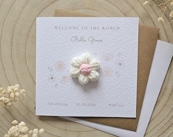 Può includere: Un biglietto bianco con il testo "Benvenuta al mondo Bella Grace" e la data "20.09.2024" stampata su di esso. Un piccolo fiore bianco all'uncinetto con un centro rosa è attaccato al biglietto.