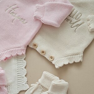 May include: A light pink knitted romper with the name Harper embroidered in white, alongside a cream knitted romper featuring the text Hello World. Both garments have wooden buttons and scalloped edges, accompanied by cream knitted baby booties.