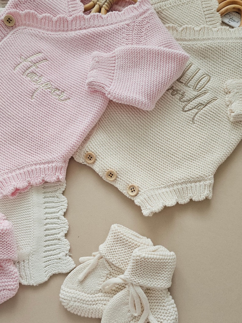 May include: A light pink knitted romper with the name Harper embroidered in white, alongside a cream knitted romper featuring the text Hello World. Both garments have wooden buttons and scalloped edges, accompanied by cream knitted baby booties.
