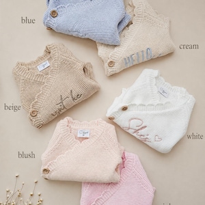 May include: A collection of six knitted wrap jumpers in blue, cream, beige, white, blush, and pink. Each jumper features a V-neckline, a single wooden button closure, and ribbed trim. Some jumpers include embroidered text like HELLO, She's, and hello.