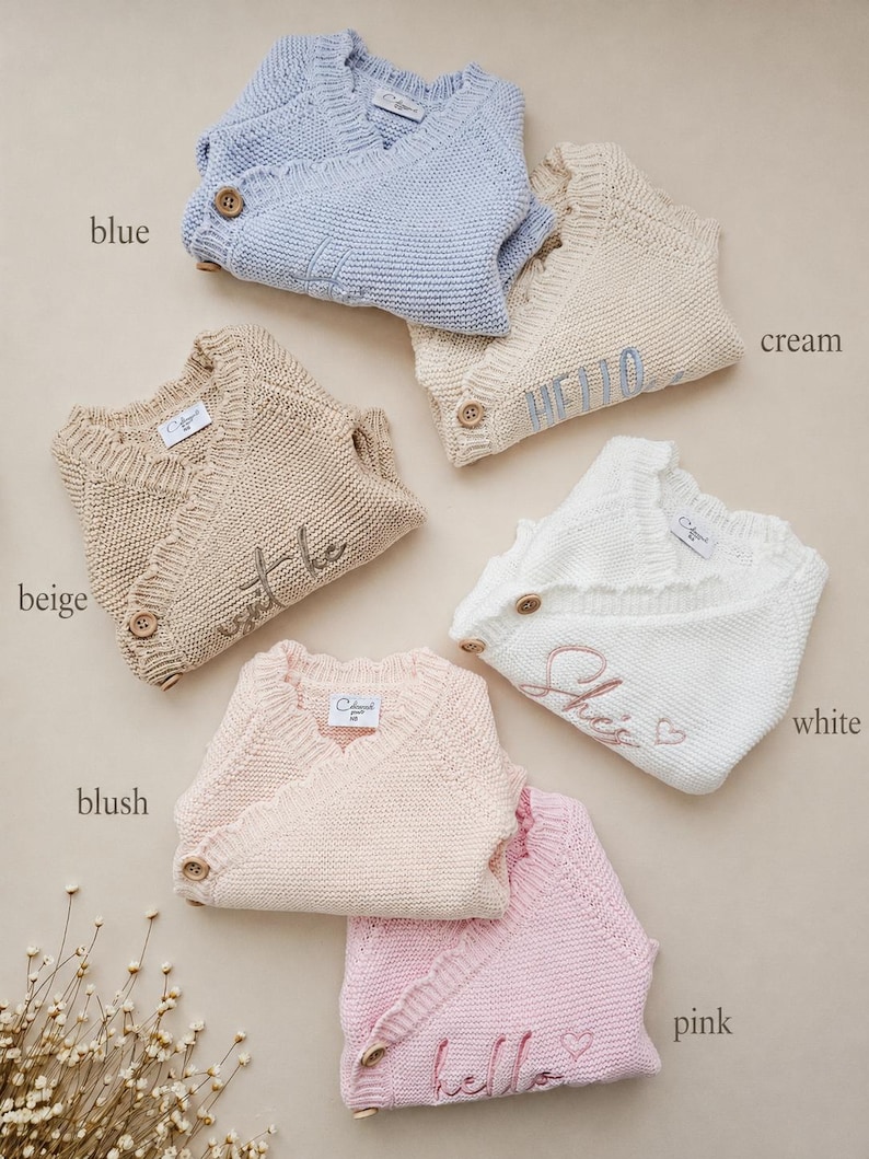 May include: A collection of six knitted wrap jumpers in blue, cream, beige, white, blush, and pink. Each jumper features a V-neckline, a single wooden button closure, and ribbed trim. Some jumpers include embroidered text like HELLO, She's, and hello.