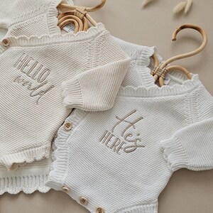 Puede incluir: Dos peleles de punto para beb&eacute;, uno crema y otro blanco, colgados de perchas de bamb&uacute;. El pelele crema tiene bordado "HELLO world" en la parte delantera. El blanco tiene "He's HERE". Ambos tienen botones de madera.