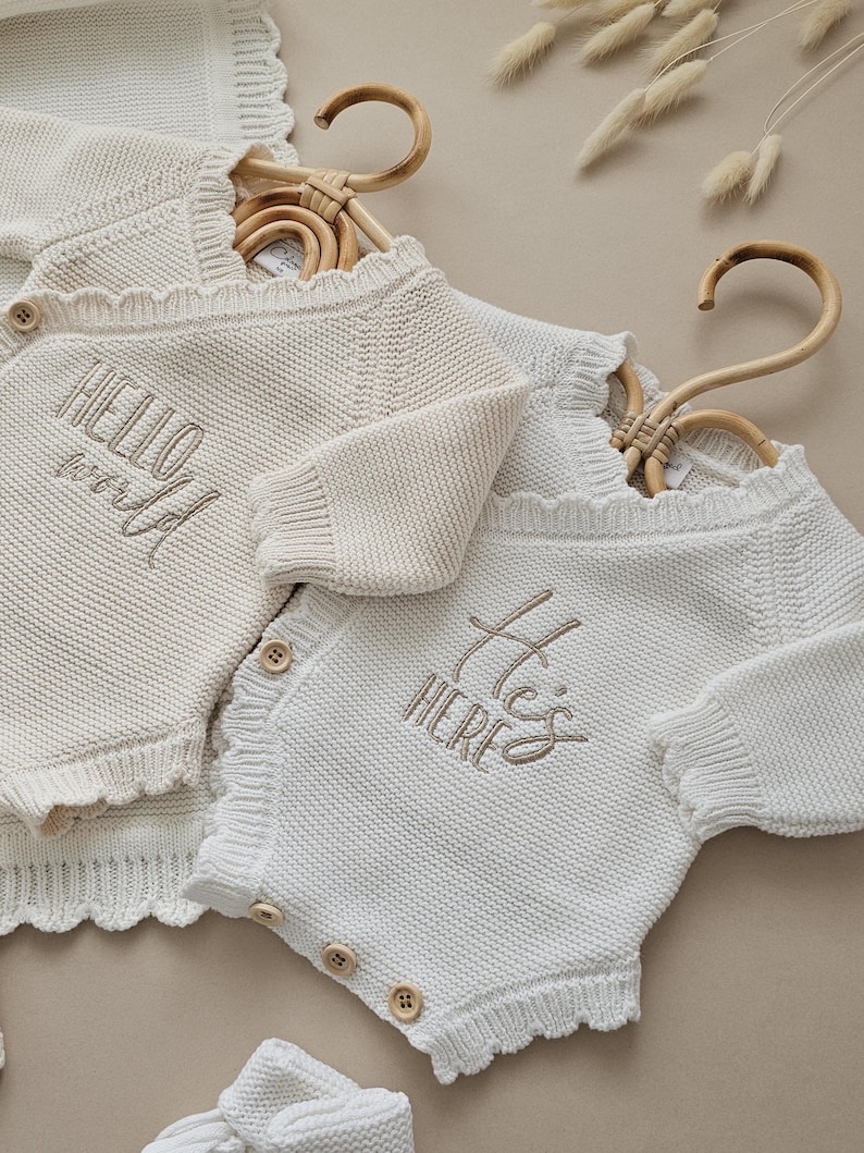 Puede incluir: Dos peleles de punto para beb&eacute;, uno crema y otro blanco, colgados de perchas de bamb&uacute;. El pelele crema tiene bordado "HELLO world" en la parte delantera. El blanco tiene "He's HERE". Ambos tienen botones de madera.