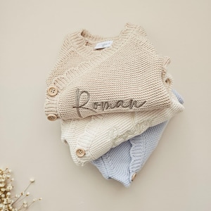 May include: A stack of three knitted jumpers in beige, cream, and light blue. The top beige jumper features the embroidered name Roman in dark thread and two wooden buttons on the side. Each jumper has a ribbed neckline and knit texture.