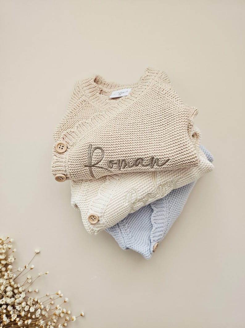 May include: A stack of three knitted jumpers in beige, cream, and light blue. The top beige jumper features the embroidered name Roman in dark thread and two wooden buttons on the side. Each jumper has a ribbed neckline and knit texture.