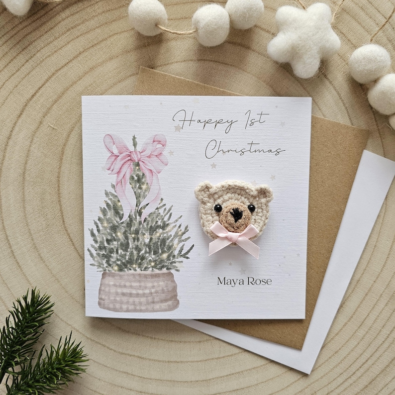 May include: A Christmas card featuring a watercolour Christmas tree and a crocheted teddy bear. The card reads "Happy 1st Christmas" and "Maya Rose". The bear has a pink bow. The card is on a wooden surface.
