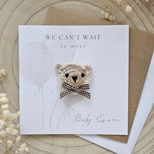 May include: A handmade baby announcement card with a crocheted teddy bear. The card reads "WE CAN'T WAIT to meet" and includes the name "Baby Evans". The bear has a checkered bow.