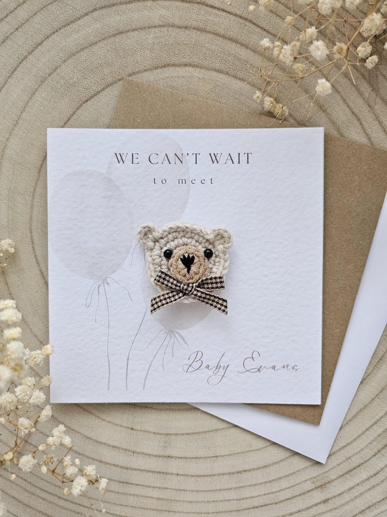 May include: A handmade baby announcement card with a crocheted teddy bear. The card reads "WE CAN'T WAIT to meet" and includes the name "Baby Evans". The bear has a checkered bow.