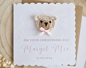May include: A white card with a scalloped edge features a crocheted bear with a pink bow. The card reads "On your christening day Margot Mia 21.07.2024".