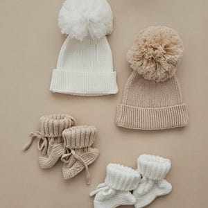 May include: Two knitted baby hats with large pom-poms, one white and one beige, are displayed alongside two pairs of matching booties. The booties have a folded cuff and tie closure. The items are arranged on a neutral background.