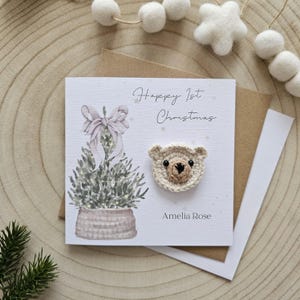 May include: A festive greeting card with a crocheted teddy bear head. The card reads "Happy 1st Christmas" and "Amelia Rose". It features a watercolour illustration of a Christmas tree with a bow.