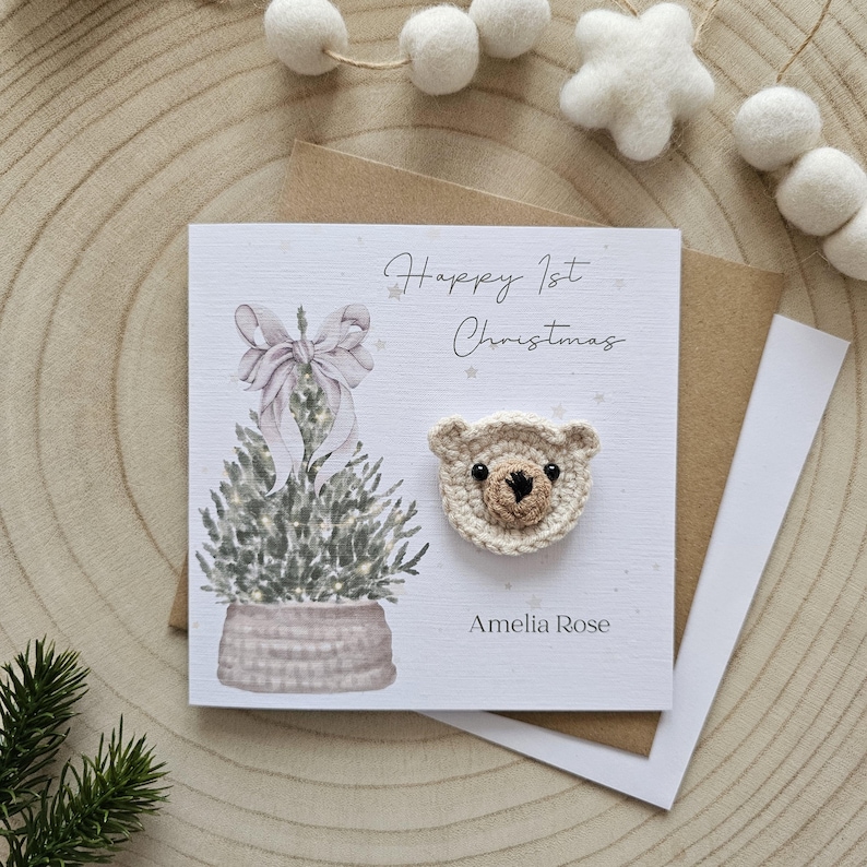 May include: A festive greeting card with a crocheted teddy bear head. The card reads "Happy 1st Christmas" and "Amelia Rose". It features a watercolour illustration of a Christmas tree with a bow.