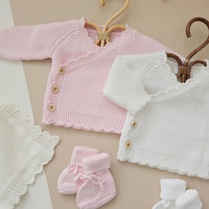 Puede incluir: Una colecci&oacute;n de ropa de beb&eacute; en rosa y blanco. La imagen presenta dos c&aacute;rdigans cruzados de punto, una manta y dos pares de patucos. Los c&aacute;rdigans tienen bordes festoneados y botones de madera. Los patucos tienen un lazo.