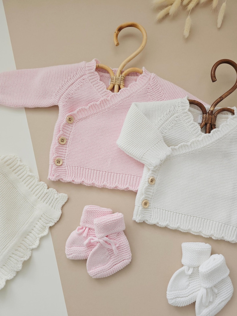 Puede incluir: Una colecci&oacute;n de ropa de beb&eacute; en rosa y blanco. La imagen presenta dos c&aacute;rdigans cruzados de punto, una manta y dos pares de patucos. Los c&aacute;rdigans tienen bordes festoneados y botones de madera. Los patucos tienen un lazo.