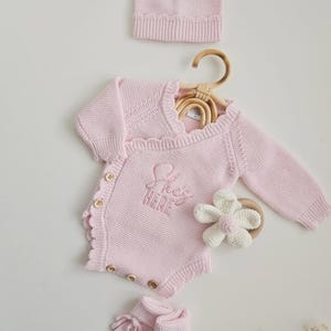 May include: A light pink knitted baby set including a long-sleeve wrap-style bodysuit with wooden buttons and the embroidered text 'She's HERE', a matching beanie, and booties. A white crocheted flower rattle with a wooden ring is included.