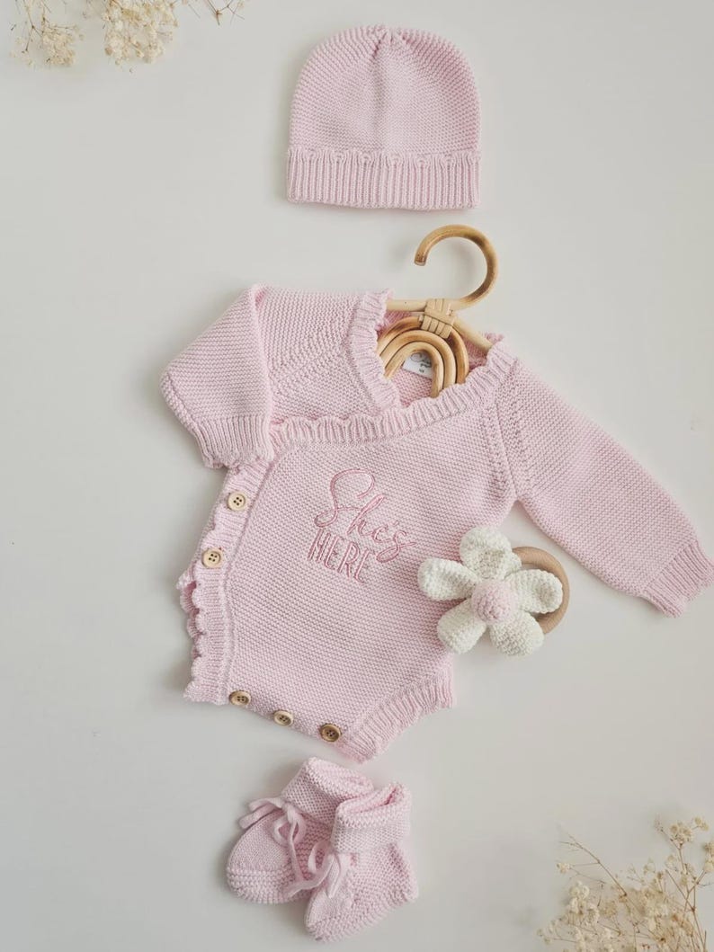 May include: A light pink knitted baby set including a long-sleeve wrap-style bodysuit with wooden buttons and the embroidered text 'She's HERE', a matching beanie, and booties. A white crocheted flower rattle with a wooden ring is included.