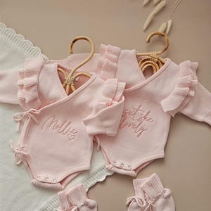 May include: Two pink baby rompers with ruffle sleeves and the embroidered text "Mollie" and "Isn't she lovely". The rompers are displayed on wooden hangers, with matching booties. The background is a neutral beige.