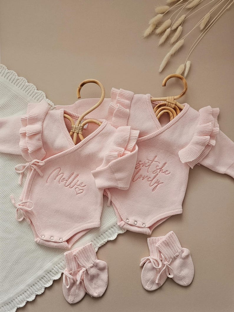 May include: Two pink baby rompers with ruffle sleeves and the embroidered text "Mollie" and "Isn't she lovely". The rompers are displayed on wooden hangers, with matching booties. The background is a neutral beige.