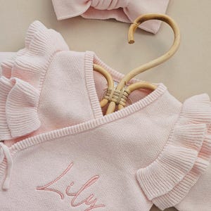 May include: A light pink baby outfit set, including a wrap-style sweater with ruffled sleeves and a matching headband. The sweater is embroidered with the name "Lily" in a delicate script. The set is displayed on a wooden hanger.