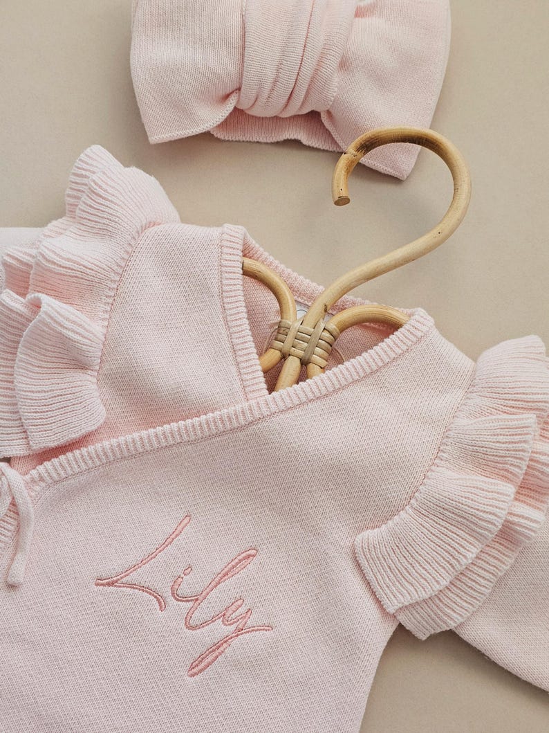 May include: A light pink baby outfit set, including a wrap-style sweater with ruffled sleeves and a matching headband. The sweater is embroidered with the name "Lily" in a delicate script. The set is displayed on a wooden hanger.