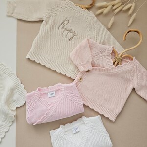 Puede incluir: Una variedad de ropa de beb&eacute; de punto en crema, rosa y blanco. La imagen presenta un su&eacute;ter color crema con el nombre "Poppy" bordado, junto con c&aacute;rdigans rosas y mantas blancas. Los art&iacute;culos est&aacute;n hechos de tejido de punto.