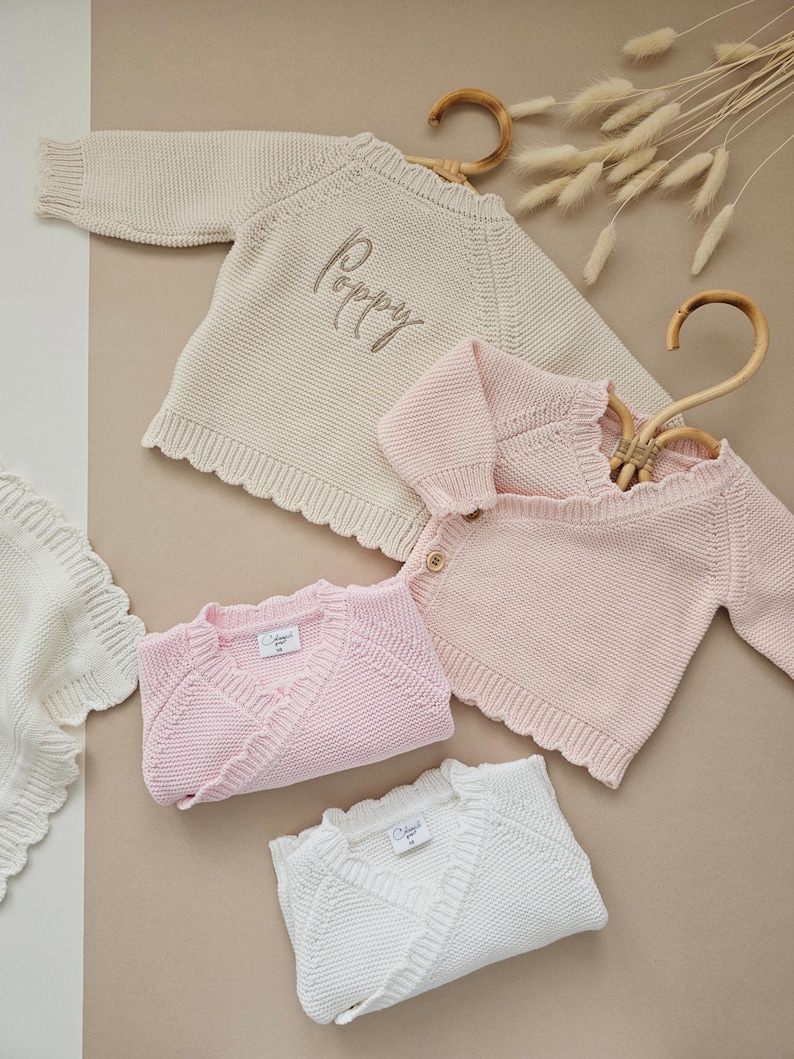 Puede incluir: Una variedad de ropa de beb&eacute; de punto en crema, rosa y blanco. La imagen presenta un su&eacute;ter color crema con el nombre "Poppy" bordado, junto con c&aacute;rdigans rosas y mantas blancas. Los art&iacute;culos est&aacute;n hechos de tejido de punto.