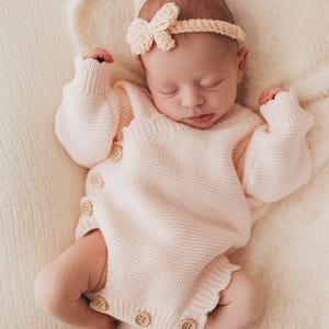 May include: A pale pink knitted long-sleeve romper featuring a side-button closure with four visible light-coloured wooden buttons and a matching crochet headband with a small bow detail, worn by a sleeping infant on a soft cream-coloured fabric.