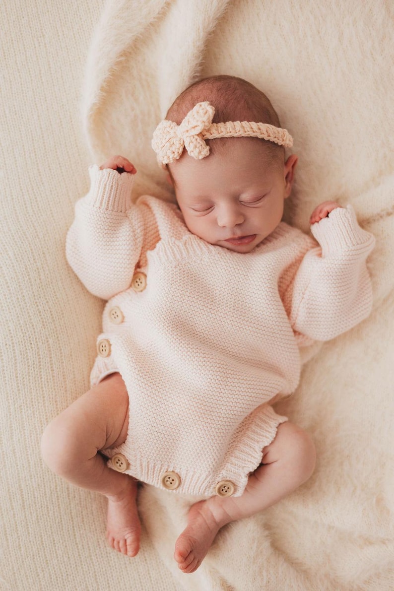 May include: A pale pink knitted long-sleeve romper featuring a side-button closure with four visible light-coloured wooden buttons and a matching crochet headband with a small bow detail, worn by a sleeping infant on a soft cream-coloured fabric.