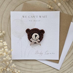 May include: A greeting card with the text "WE CAN'T WAIT to meet" and the name "Baby Evans". The card features a brown crocheted teddy bear with a beige bow. The card is on a wooden surface with dried flowers.