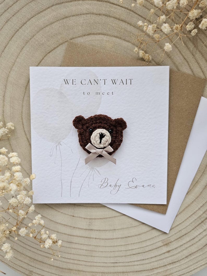May include: A greeting card with the text "WE CAN'T WAIT to meet" and the name "Baby Evans". The card features a brown crocheted teddy bear with a beige bow. The card is on a wooden surface with dried flowers.
