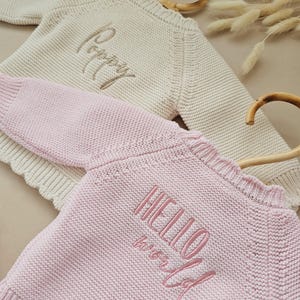 Puede incluir: Dos su&eacute;teres de beb&eacute; tejidos, uno color crema con "Poppy" bordado en dorado y otro rosa con "HELLO world" bordado en rosa. Ambos su&eacute;teres tienen un escote festoneado y se muestran en perchas de madera.