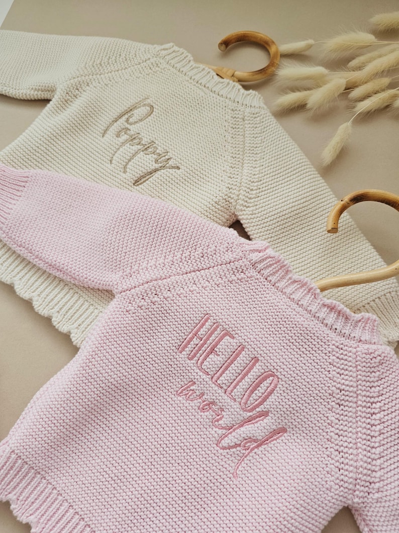 Puede incluir: Dos su&eacute;teres de beb&eacute; tejidos, uno color crema con "Poppy" bordado en dorado y otro rosa con "HELLO world" bordado en rosa. Ambos su&eacute;teres tienen un escote festoneado y se muestran en perchas de madera.
