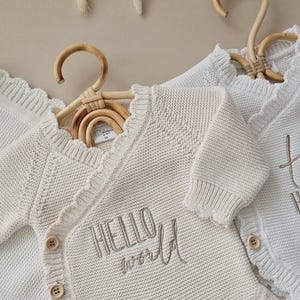 Puede incluir: Rebeca de beb&eacute; color crema con borde festoneado y el texto bordado "HELLO world". La rebeca tiene botones de madera y se muestra en una percha de madera. La prenda de punto es suave y acogedora.