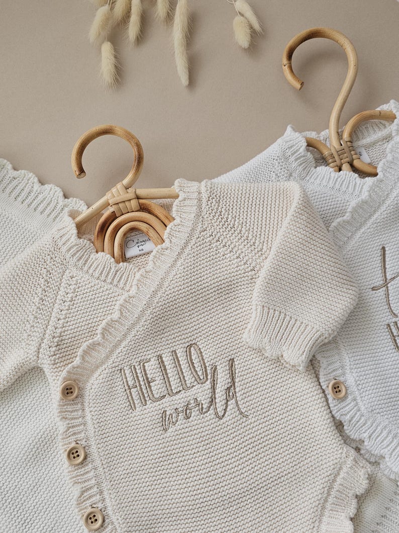 Puede incluir: Rebeca de beb&eacute; color crema con borde festoneado y el texto bordado "HELLO world". La rebeca tiene botones de madera y se muestra en una percha de madera. La prenda de punto es suave y acogedora.
