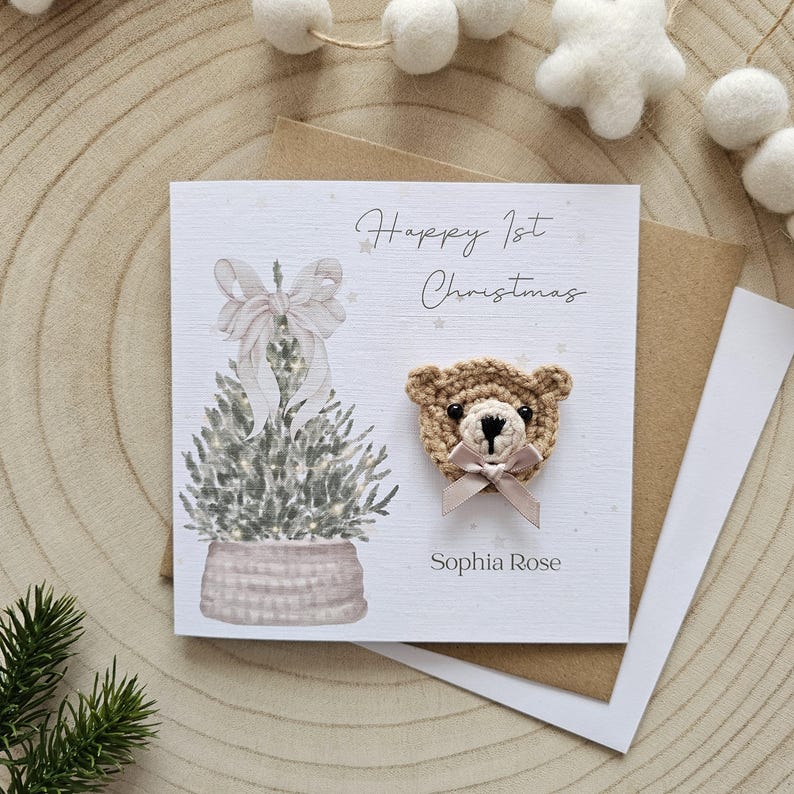 May include: A Christmas card featuring a crocheted teddy bear and a watercolour illustration of a Christmas tree. The card reads "Happy 1st Christmas" and "Sophia Rose". The card is on a wooden surface.