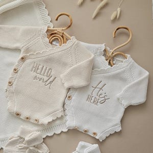 Puede incluir: Conjunto de ropa de beb&eacute; color crema, que incluye dos bodies de punto con las frases "Hello World" y "He's Here" bordadas. El conjunto tambi&eacute;n incluye patucos y una manta a juego.