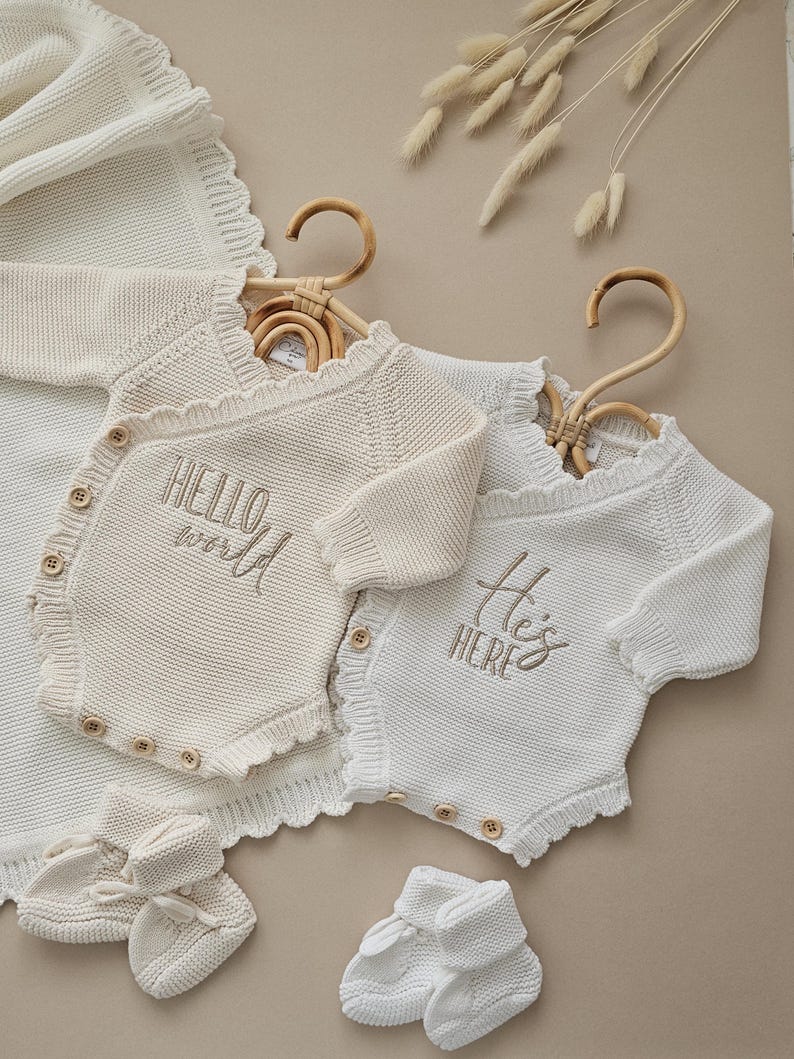 Puede incluir: Conjunto de ropa de beb&eacute; color crema, que incluye dos bodies de punto con las frases "Hello World" y "He's Here" bordadas. El conjunto tambi&eacute;n incluye patucos y una manta a juego.