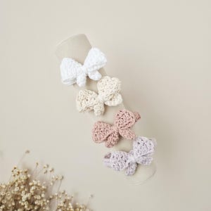 May include: Five handmade crochet hair bows in pastel shades of white, cream, dusty rose, and lavender. The bows are displayed on a beige cylindrical form, highlighting their delicate, textured design. The background is a soft, neutral tone.